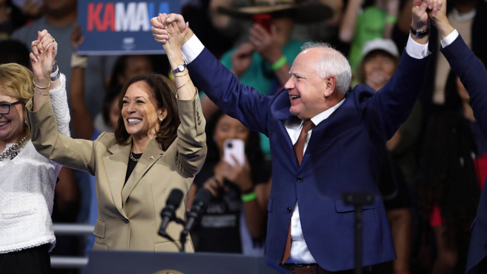 Kamala Harris and supporters celebrate together at a campaign rally, raising hands in unity with an enthusiastic crowd in the background.