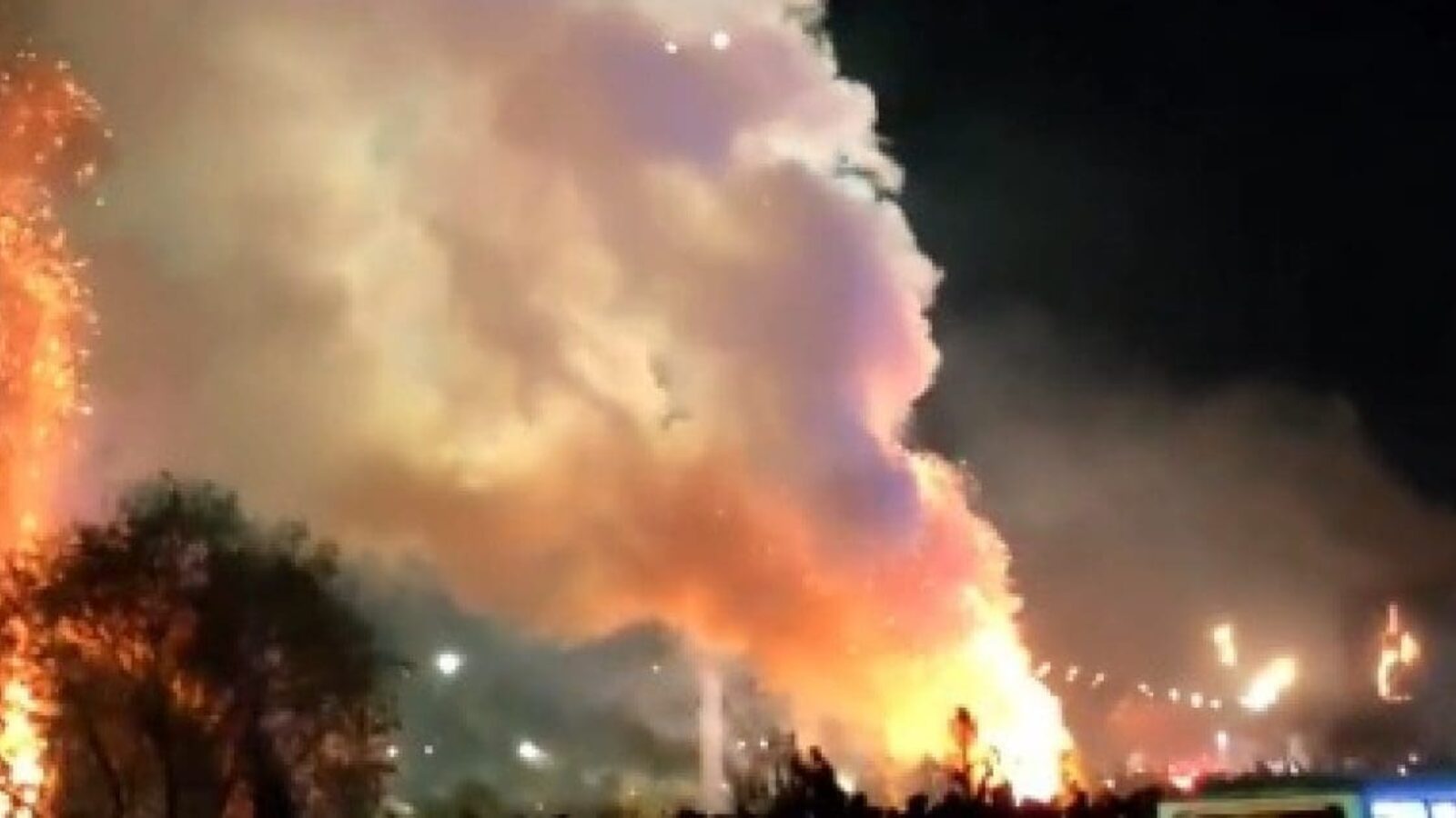 Vibrant flames and colorful smoke rise into the night sky during a fireworks display, illuminating the surrounding area with bright colors and sparks.