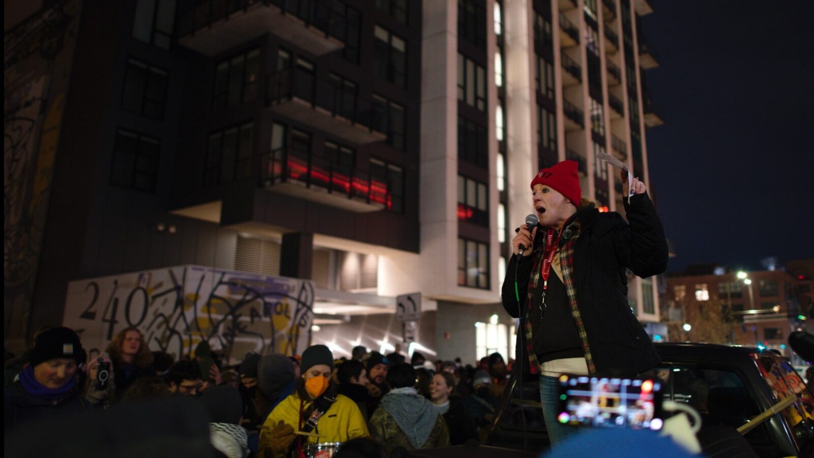 A speaker passionately addresses a crowd during a nighttime protest, with urban buildings and graffiti visible in the background.