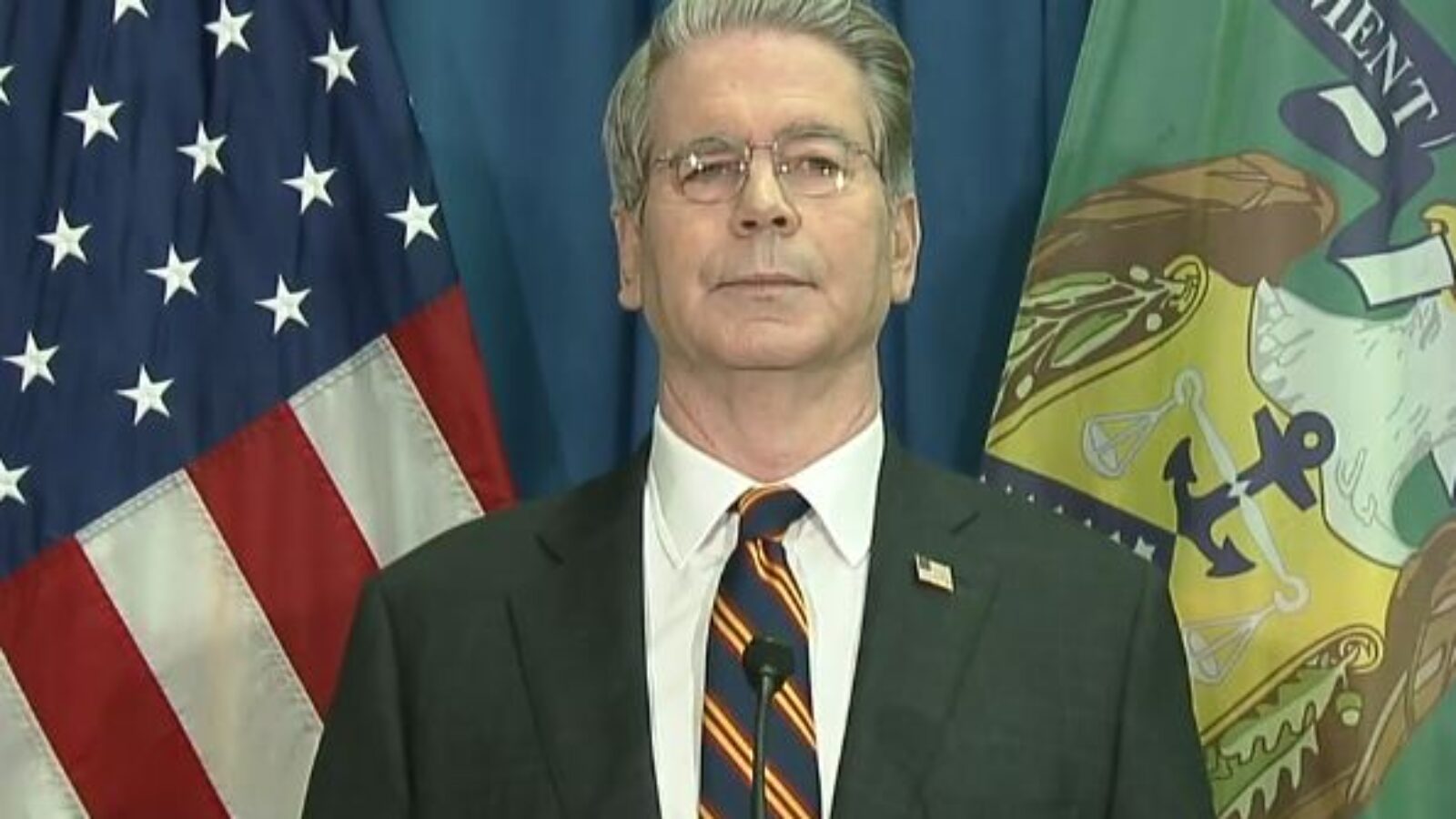 Man standing in front of American flag and department banner, delivering a statement on a podium during a press conference.