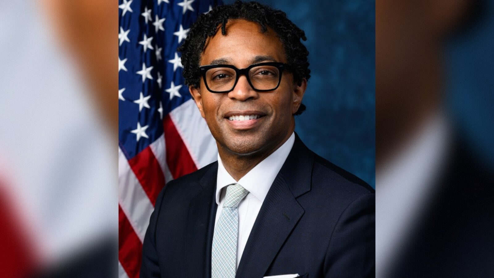 Portrait of a professional man in a suit with glasses, smiling in front of an American flag backdrop.