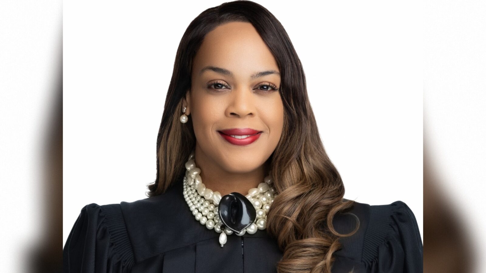 Portrait of a confident female judge wearing a black robe and pearl necklace, smiling against a plain white background.
