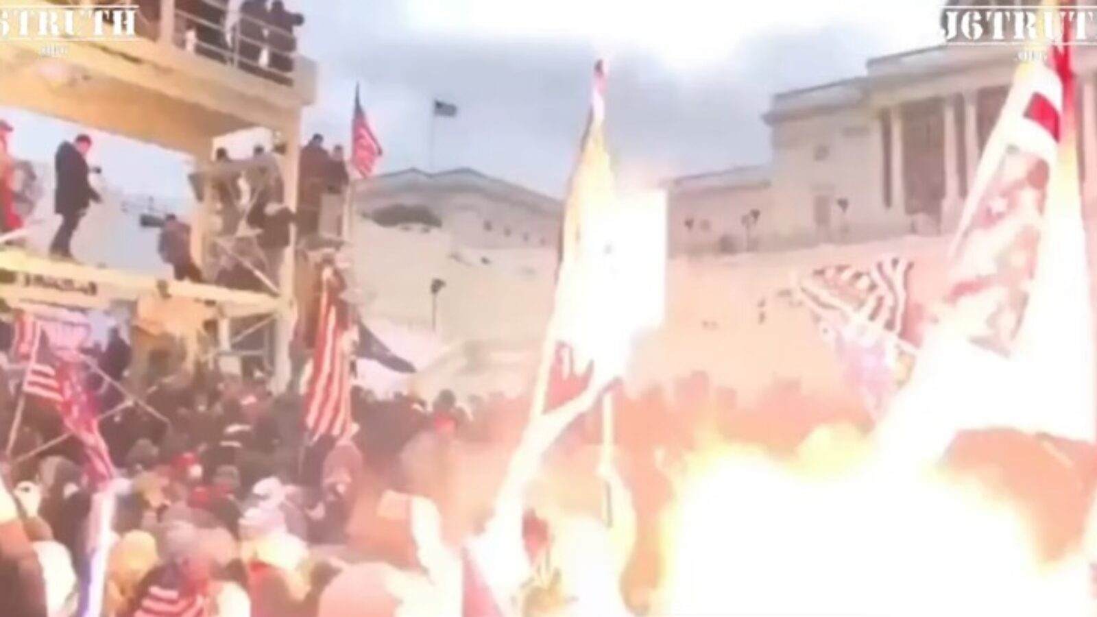 Crowd of protesters at a rally, waving flags and gathered near a building, with smoke and flames visible in the foreground.