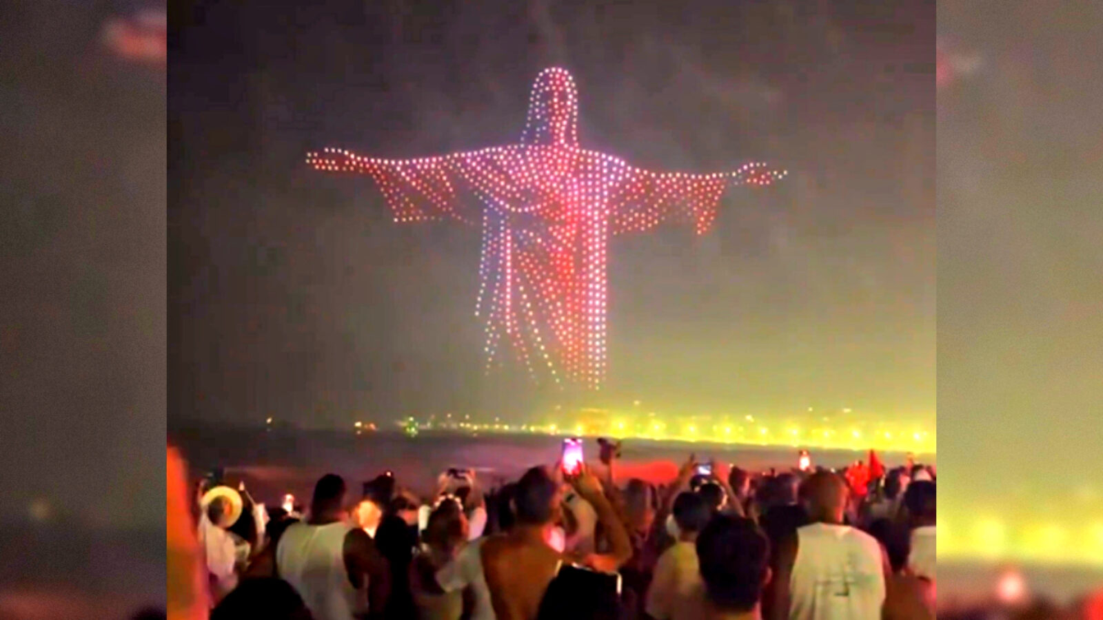 Crowd gathered on the beach watching a spectacular light display of the Christ the Redeemer statue against a night sky.