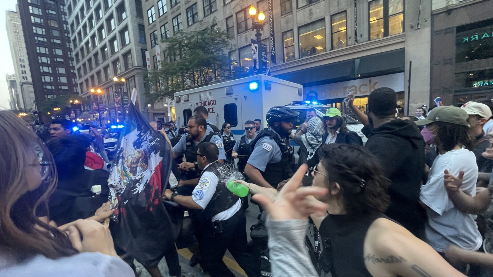 Crowd of protesters and police officers in downtown Chicago during a demonstration, with emergency vehicles and a diverse group of people showing various expressions and actions.