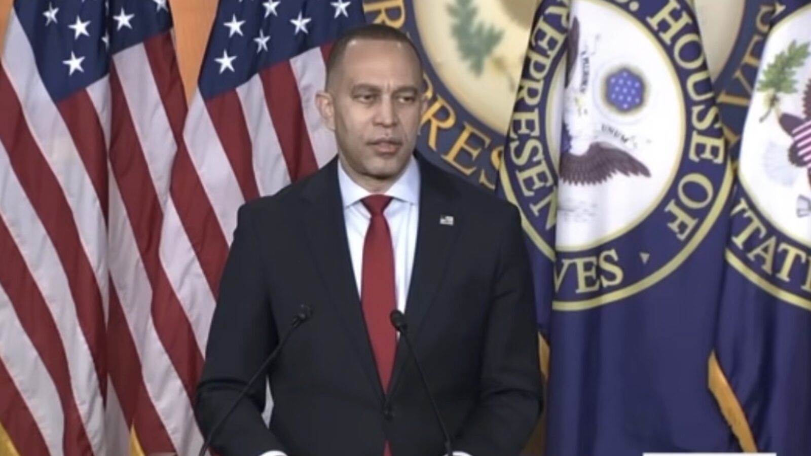 U.S. Congressman delivering a speech in front of American flags and congressional seals, emphasizing his political agenda and initiatives.