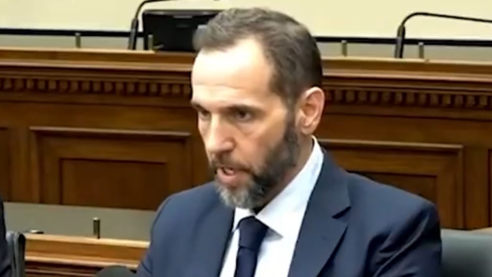 Man in a suit speaking during a formal meeting, with a wooden backdrop and empty seats visible, conveying a serious tone.