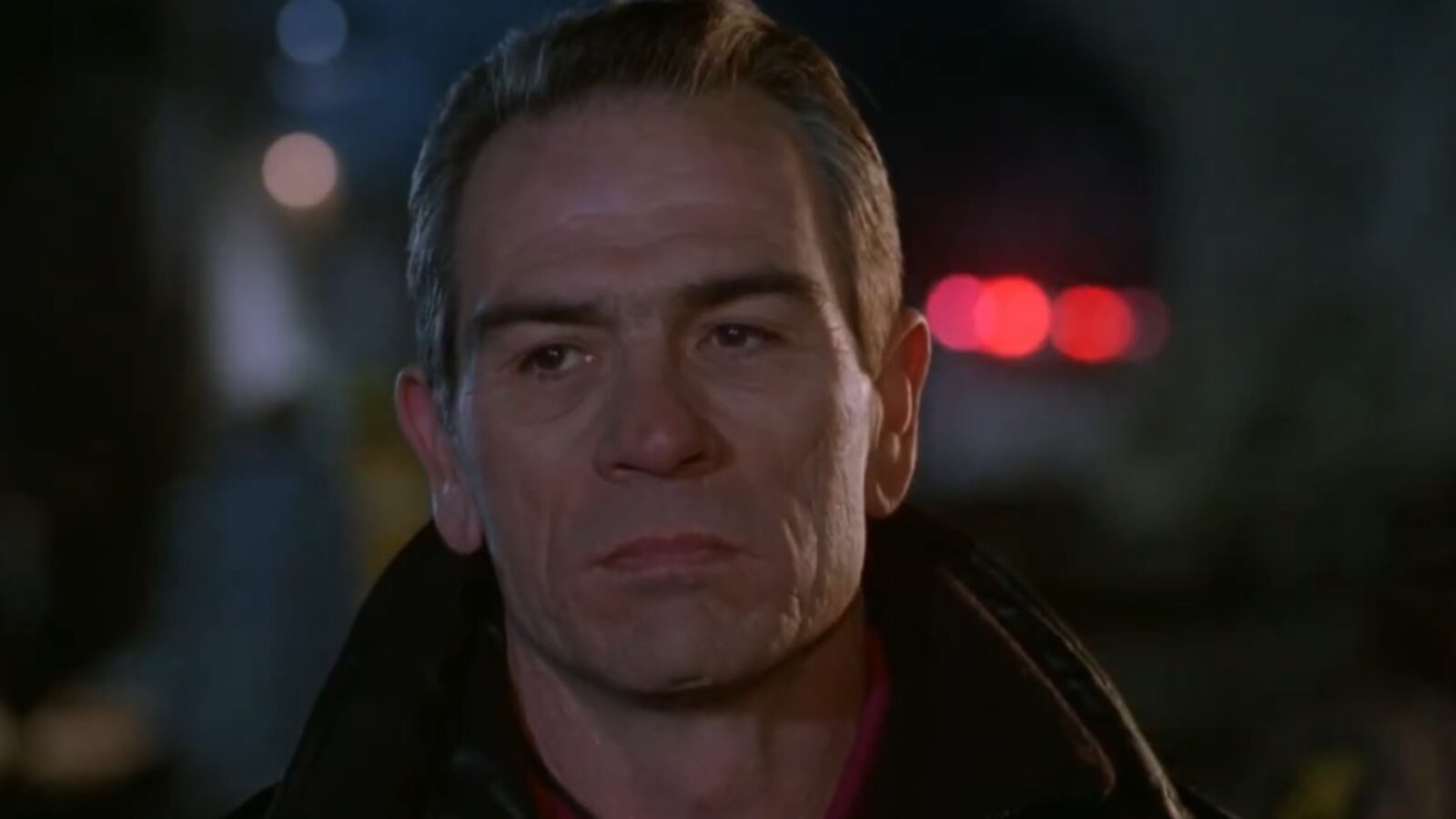 Close-up of a serious man with gray hair, wearing a black jacket, against a blurred nighttime background with red lights.
