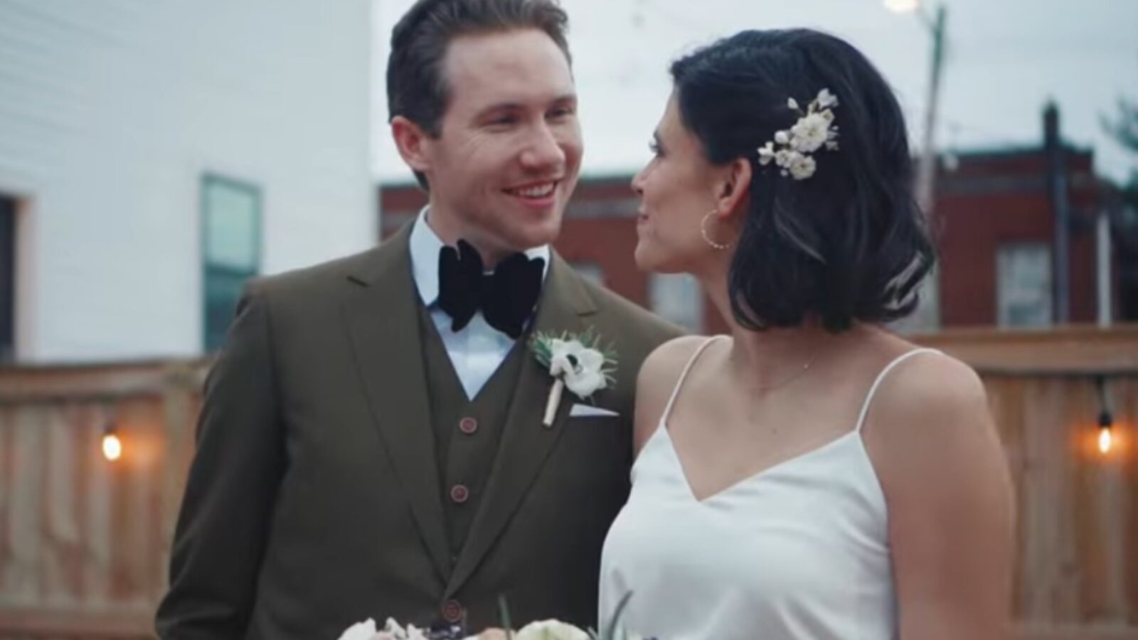 A smiling couple in formal wedding attire stands together outdoors, with the bride holding a bouquet and floral accents in her hair.