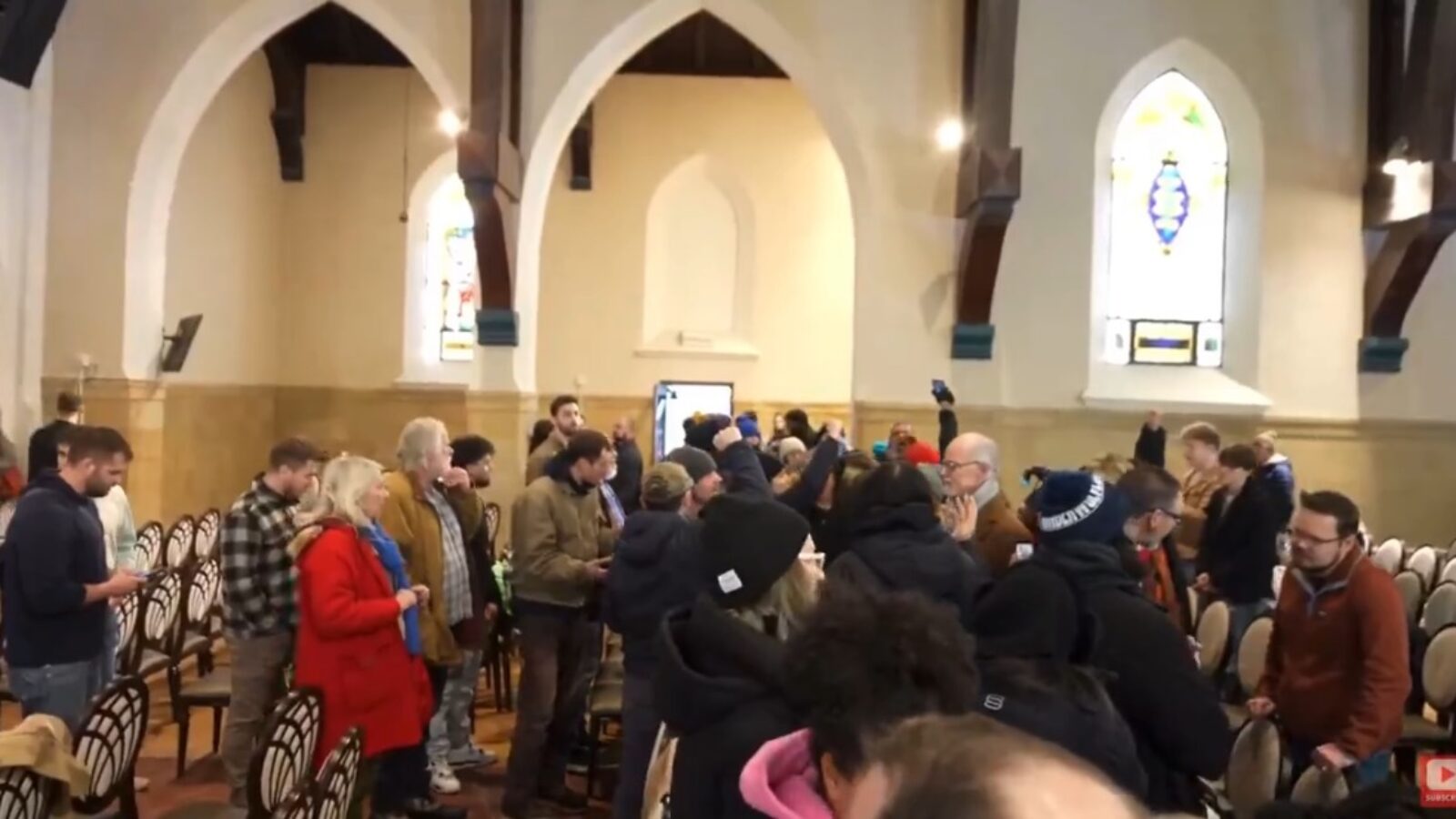 A crowd of people gathers inside a church with stained glass windows, engaging in conversation and using smartphones.