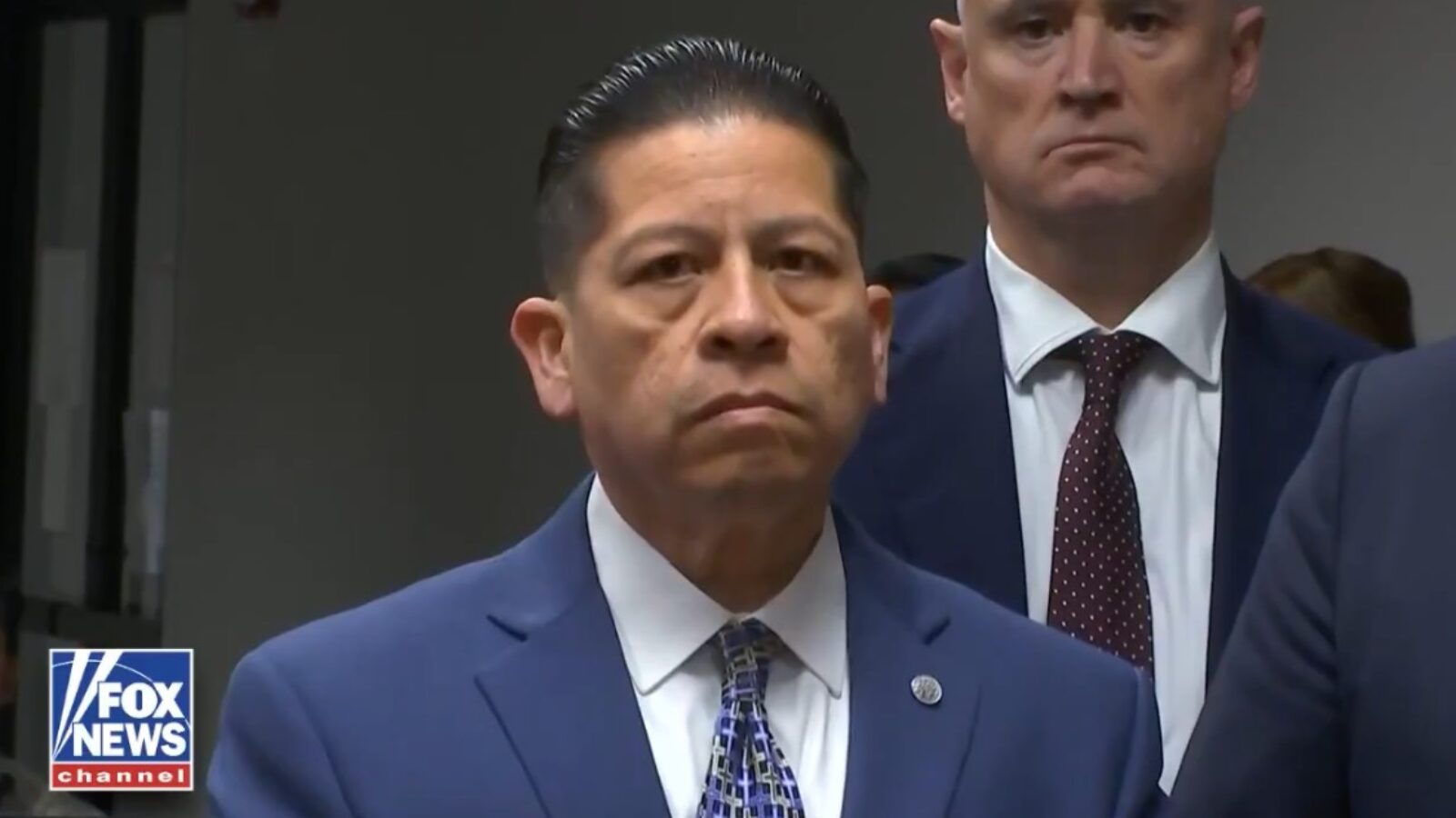 A serious-looking man in a blue suit stands in front of a group of people, conveying a sense of authority during a press event.