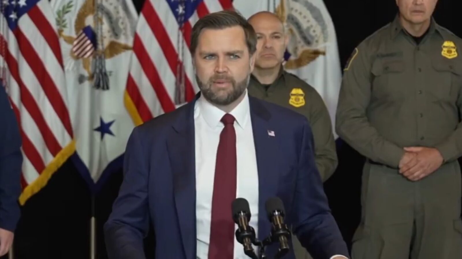Official press conference featuring a speaker in a suit addressing an audience with American flags in the background, flanked by law enforcement personnel.