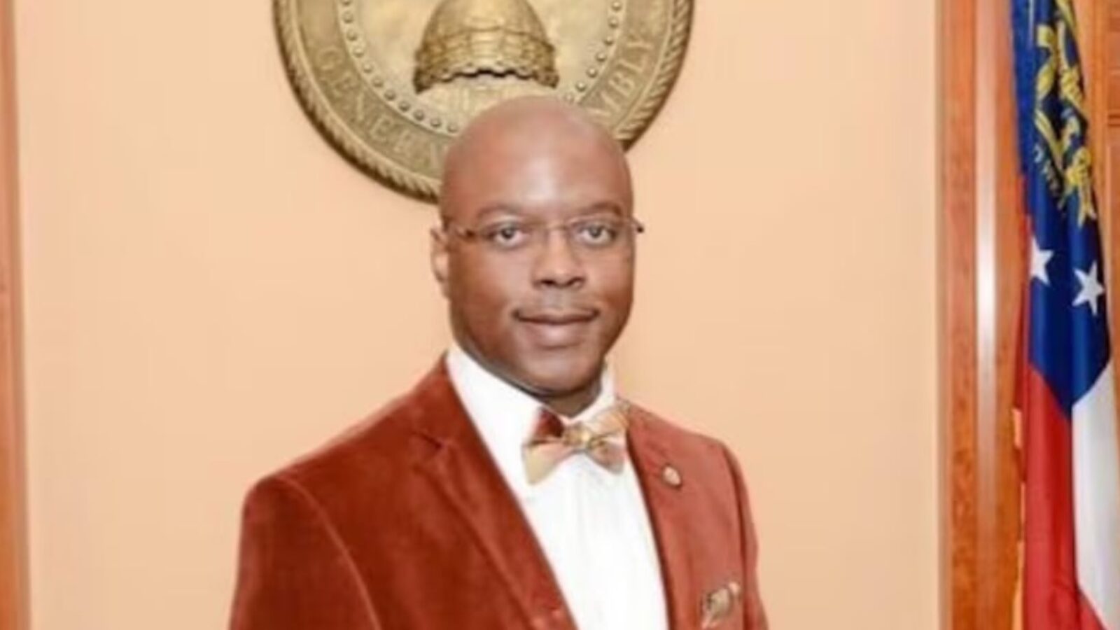 Portrait of a professional man in a brown suit with a bow tie, standing in front of a state seal and flags, conveying a formal and authoritative presence.