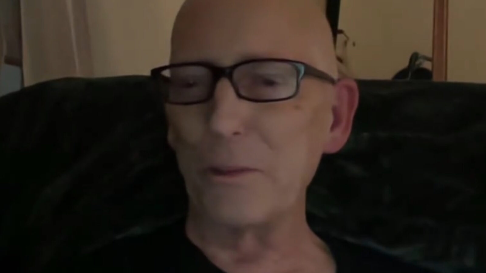 Elderly man with glasses sitting on a couch, engaged in conversation, displaying a thoughtful expression in a cozy indoor setting.