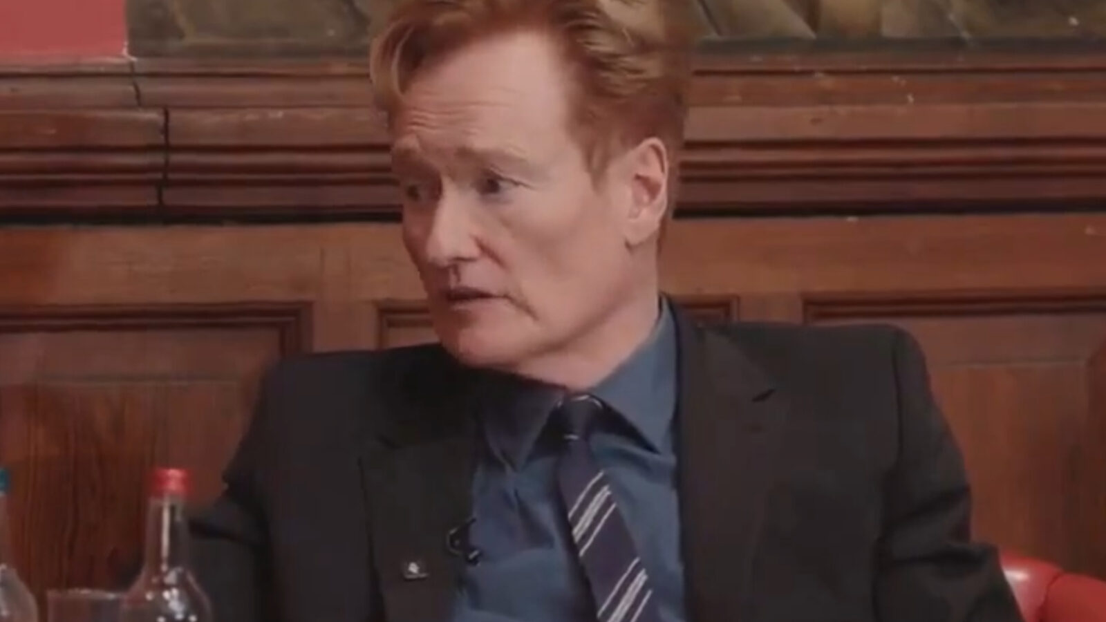 Conan O'Brien speaking during a discussion in a formal setting, wearing a suit and seated with water bottles on the table.
