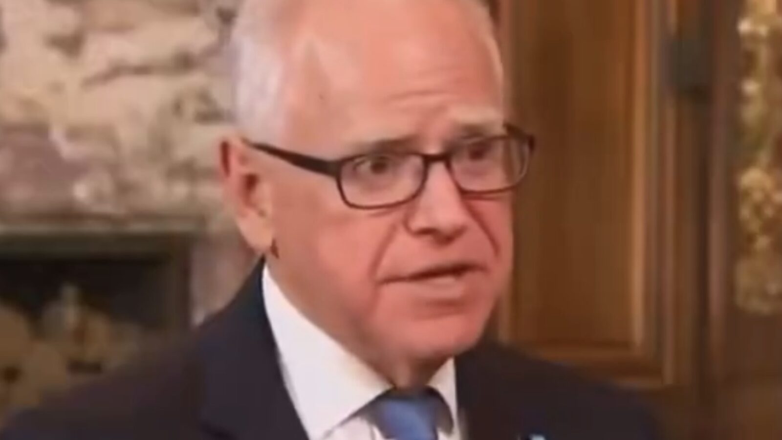 Business professional speaking in an elegant setting, wearing glasses and a suit with a blue tie, engaged in a serious conversation.