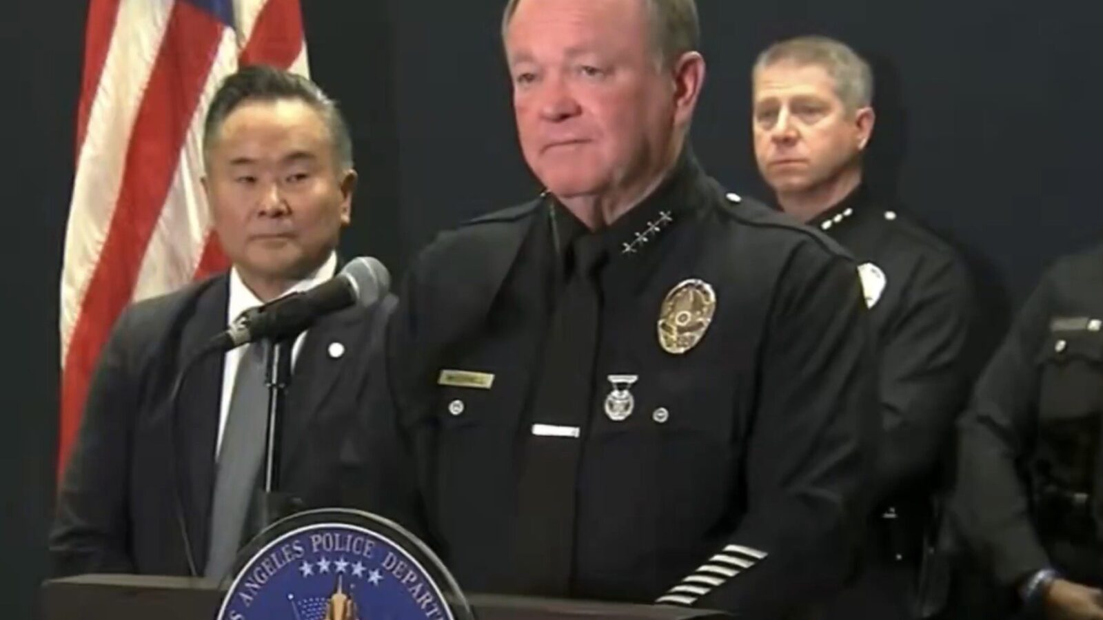 Los Angeles Police Chief addresses the media during a press conference, with officers and an official in the background, alongside the LAPD emblem and American flag.