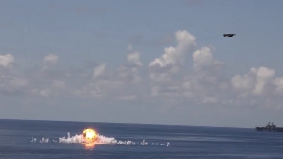 Military aircraft conducts a strike over ocean, resulting in a large explosion and water splash against a backdrop of clouds.
