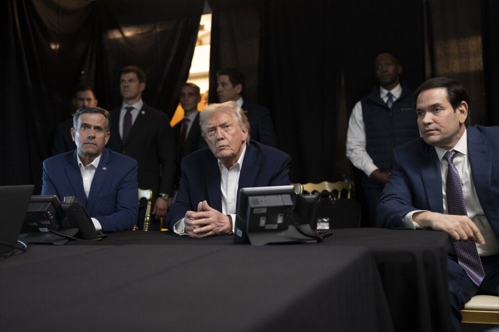 Donald Trump and two officials sit at a table with phones, engaged in a serious discussion while others stand in the background.
