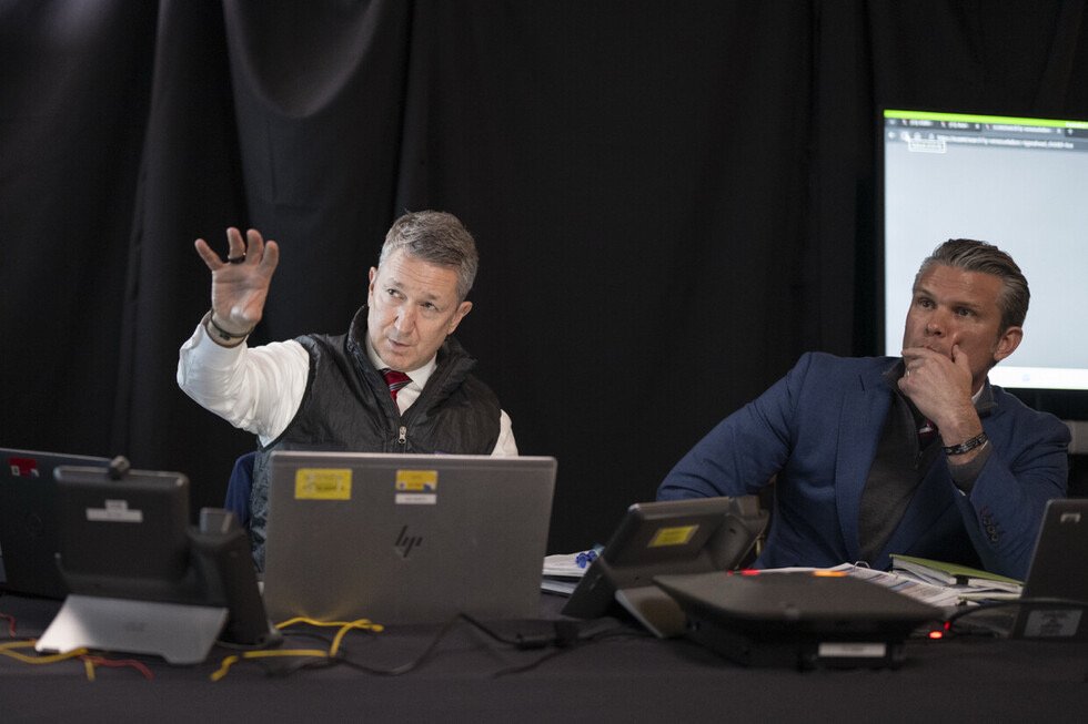 Two professionals engaged in a discussion at a conference table with multiple computers and digital screens in a dark setting.