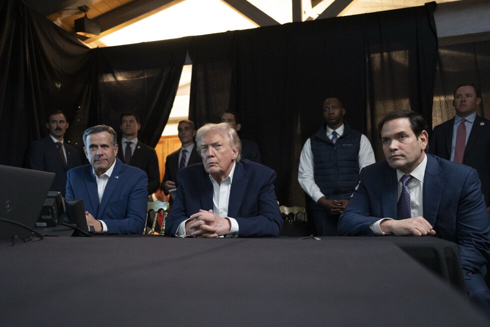 Donald Trump and two advisors seated at a table during a meeting, with security personnel in the background and a dark curtain behind them.