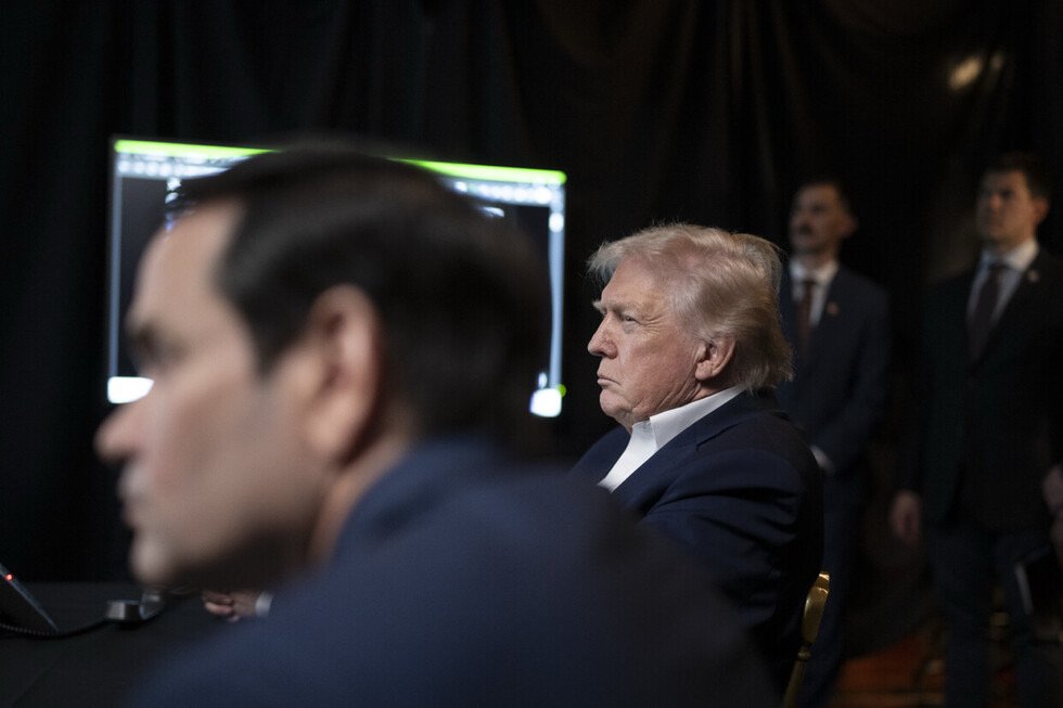 Donald Trump listens intently during a meeting, with advisors and a computer screen visible in the background.