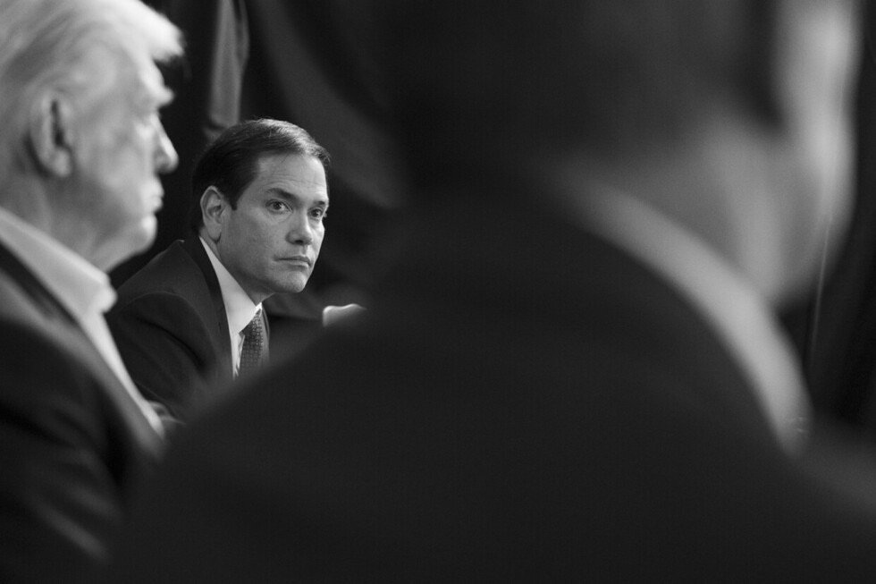 Black and white image of Marco Rubio during a discussion, with a blurred figure of Donald Trump in the foreground, highlighting political engagement.