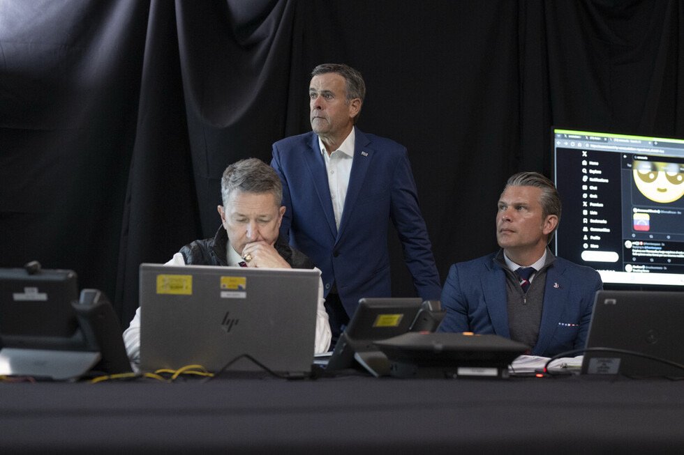 Three professionals focus on computer screens in a dimly lit control room, discussing strategy and analyzing data during a critical meeting.