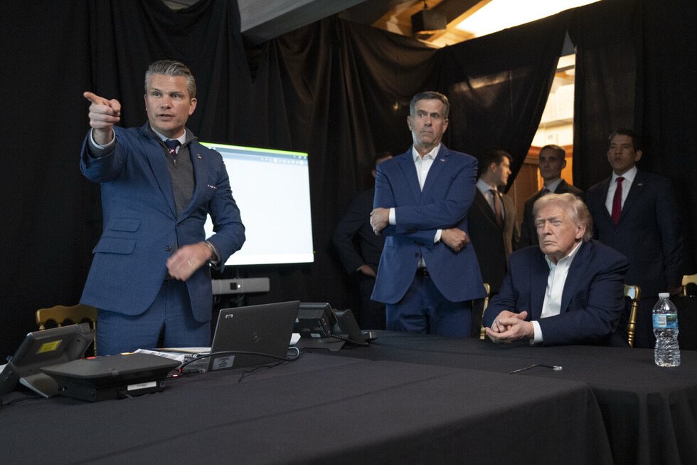 A group of men, including a seated Donald Trump, discuss strategy during a meeting, with technology and a presentation screen in the background.