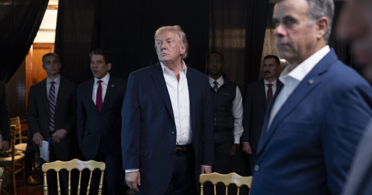 Donald Trump stands among advisors and security personnel in a dimly lit setting, conveying a moment of contemplation or preparation.