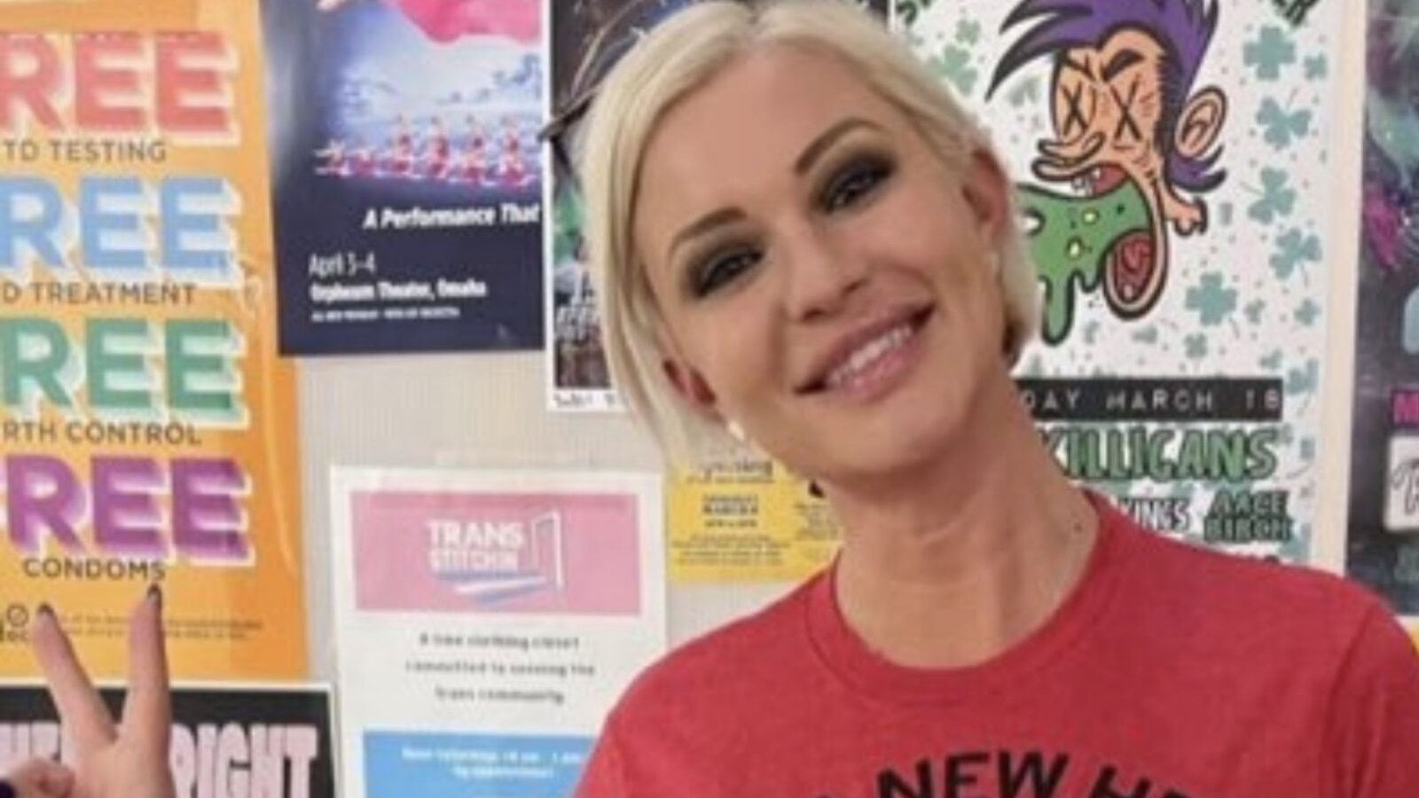 Smiling woman in a red shirt poses with peace sign in front of colorful community health awareness posters promoting free services and events.