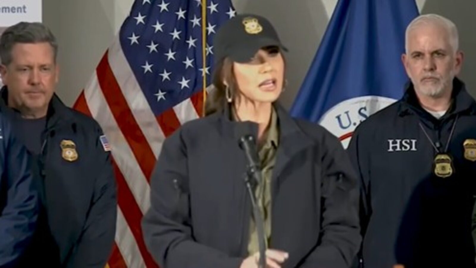 Official press conference featuring a female speaker in a cap, flanked by law enforcement officials and U.S. flags, discussing immigration enforcement.