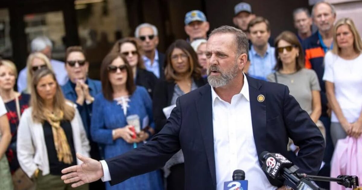 A man in a suit speaks passionately at a press conference, surrounded by a diverse group of attentive supporters and media representatives.