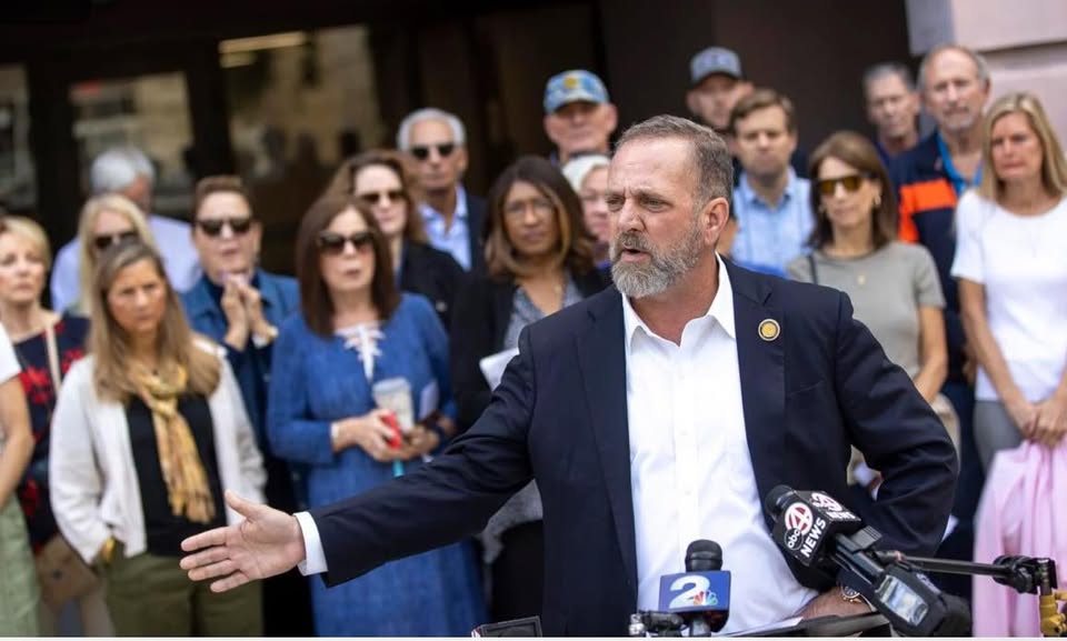 A man in a suit speaks passionately at a press conference, surrounded by a diverse group of attentive supporters and media representatives.
