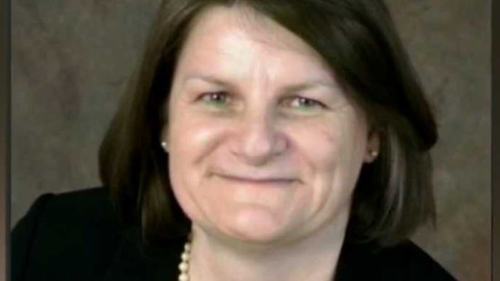 Smiling woman with shoulder-length brown hair wearing a black blazer and pearl necklace, set against a neutral background.