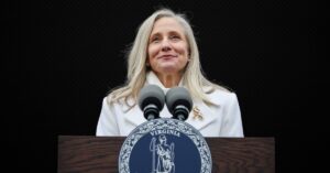 Woman in a white coat speaks at a podium featuring the Virginia state seal, highlighting a public address or event.