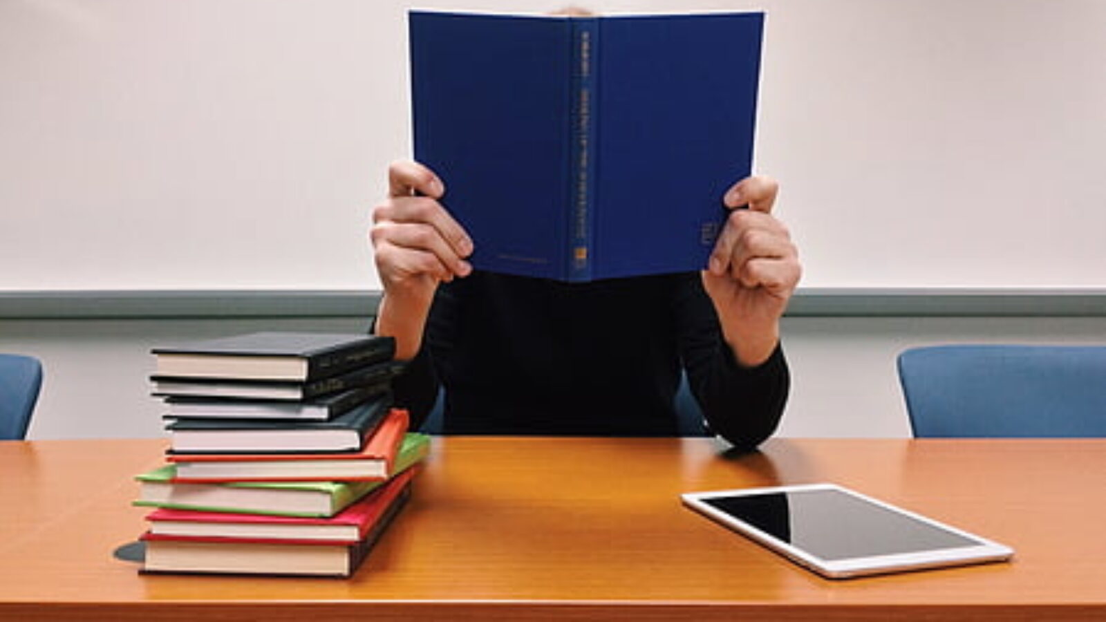Person reading a blue book at a table with a stack of colorful books and a tablet nearby.