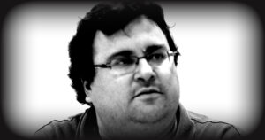 Portrait of a man with dark hair and glasses, looking thoughtfully to the side against a blurred background.