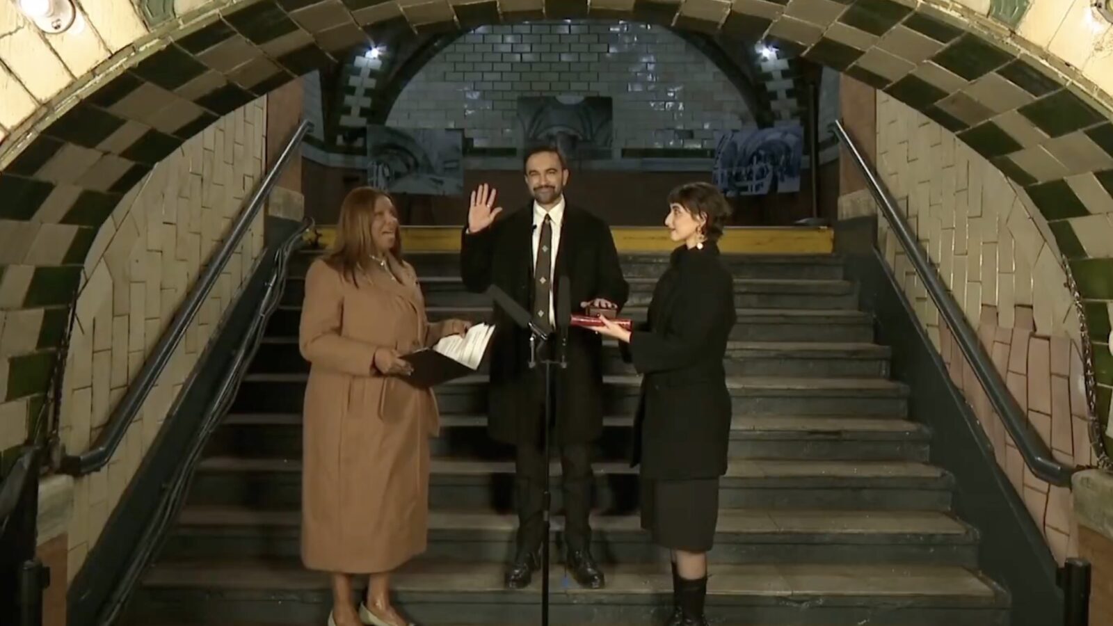 Ceremony in a subway station featuring a man taking an oath, flanked by two women holding documents, with an ornate archway in the background.