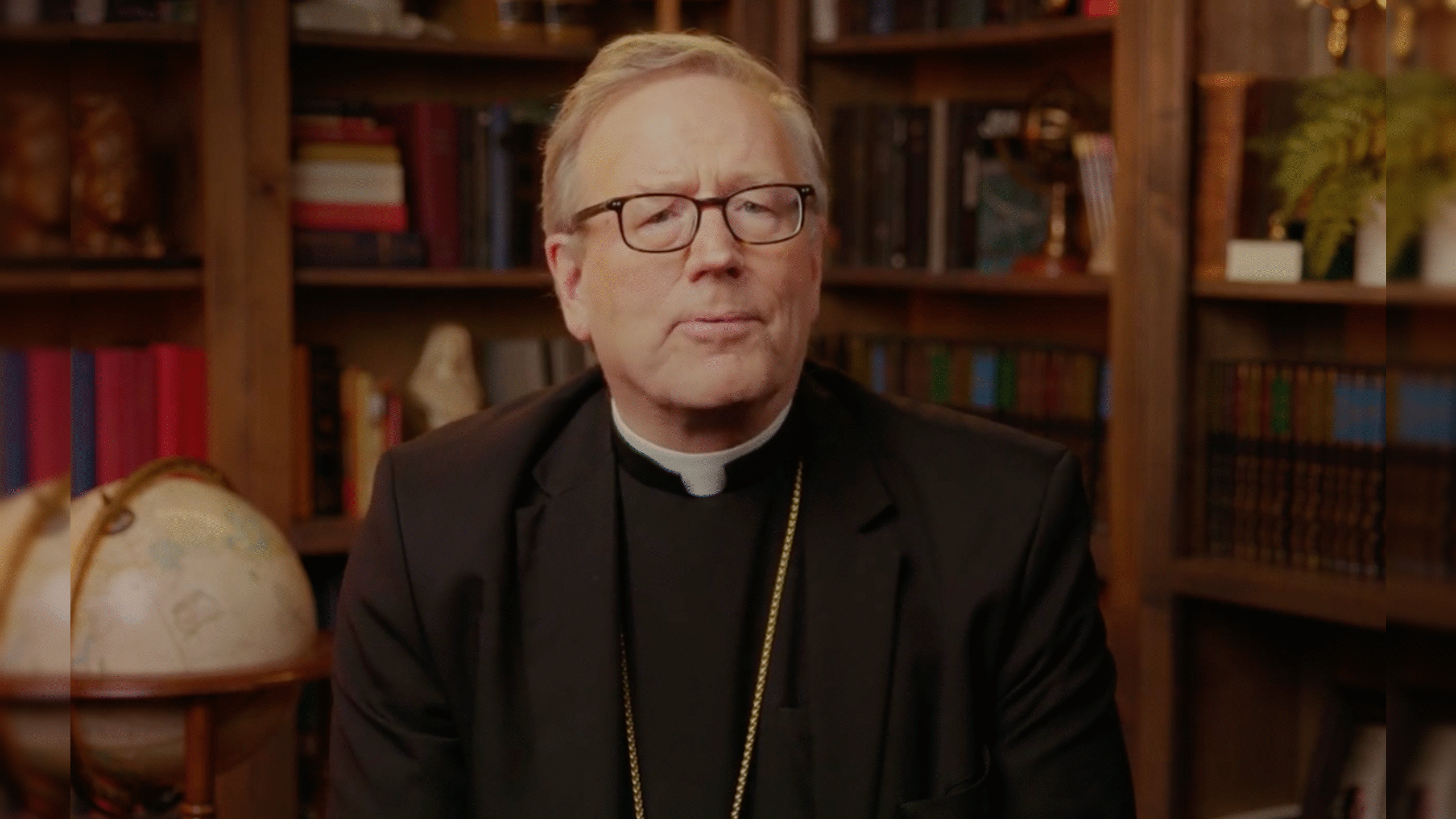 Clergyman in a black suit and glasses speaking in front of a bookshelf and globe, conveying a message with a thoughtful expression.