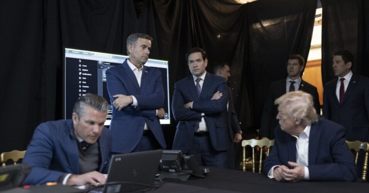A group of men, including Donald Trump, are engaged in a serious discussion around a table with a computer and a large screen displaying data.