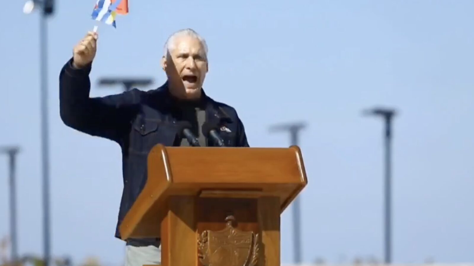 Man passionately speaking at a podium while holding a flag, set against a clear blue sky.