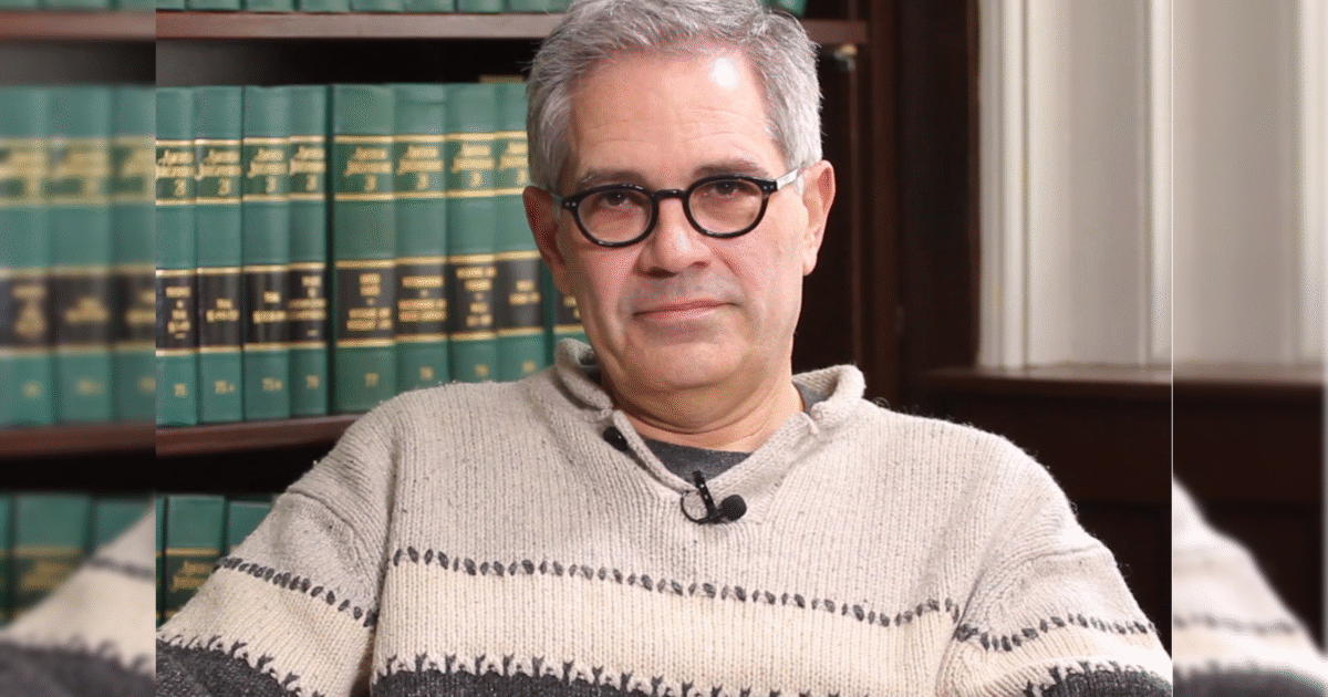 Man with gray hair and glasses sitting in front of a bookshelf filled with law books, wearing a light sweater.