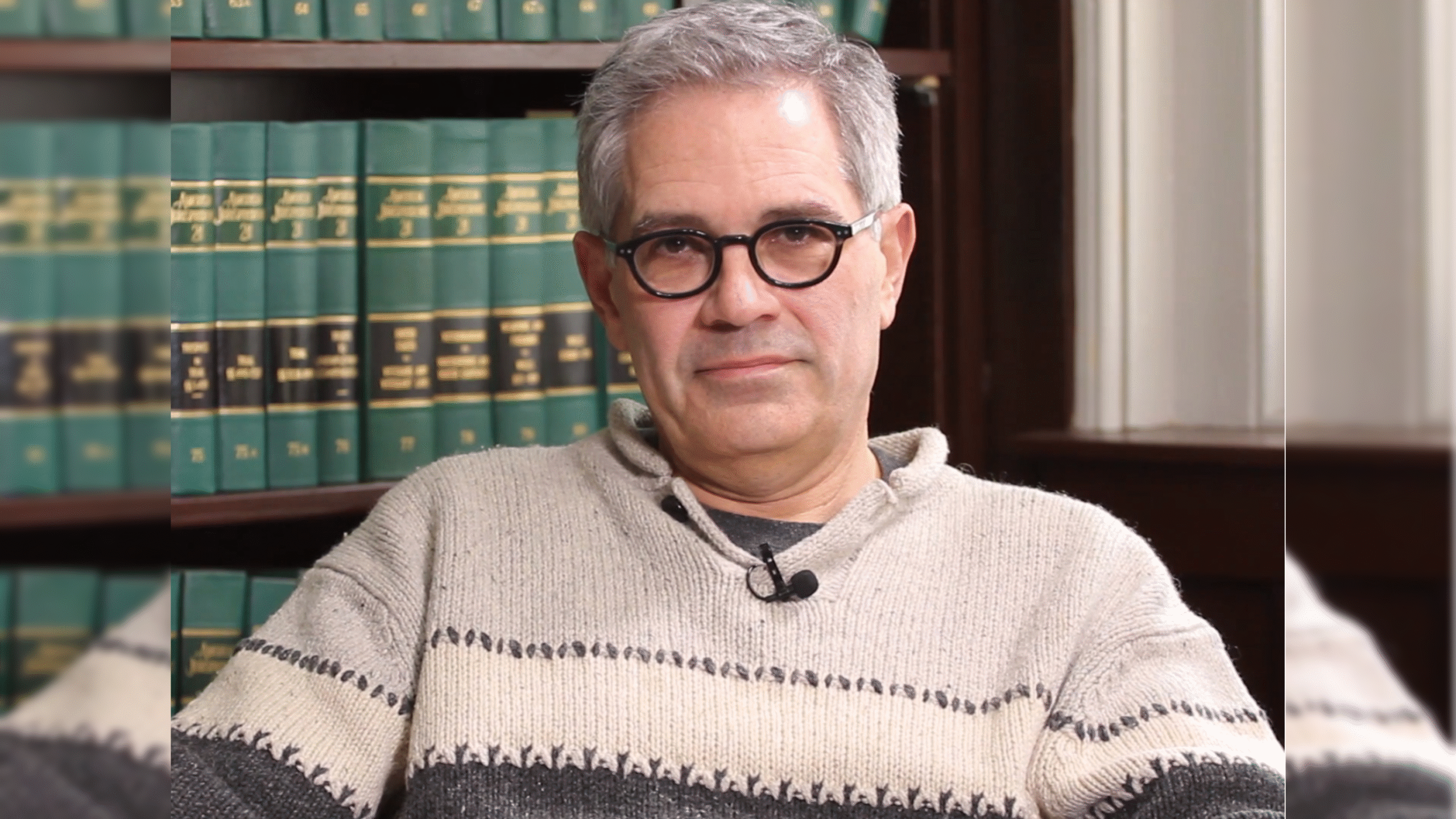 Man with gray hair and glasses sitting in front of a bookshelf filled with law books, wearing a light sweater.