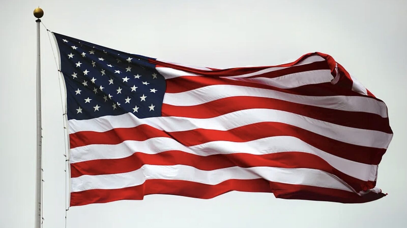 American flag waving against a gray sky, showcasing red and white stripes with blue field of stars, symbolizing patriotism and national pride.