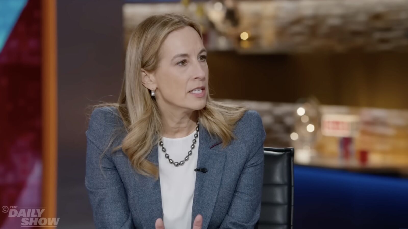 A woman with long hair, wearing a blue blazer and pearl necklace, speaks thoughtfully during an interview on a talk show set.