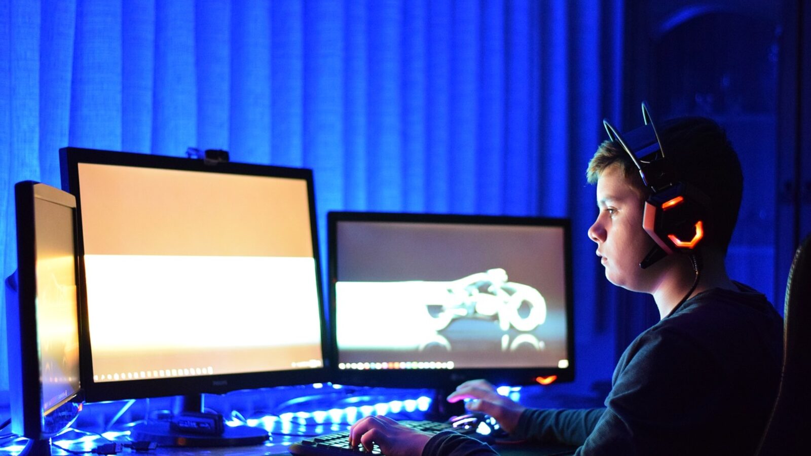 Young gamer wearing a headset, focused on dual monitors in a dimly lit room with blue ambient lighting.