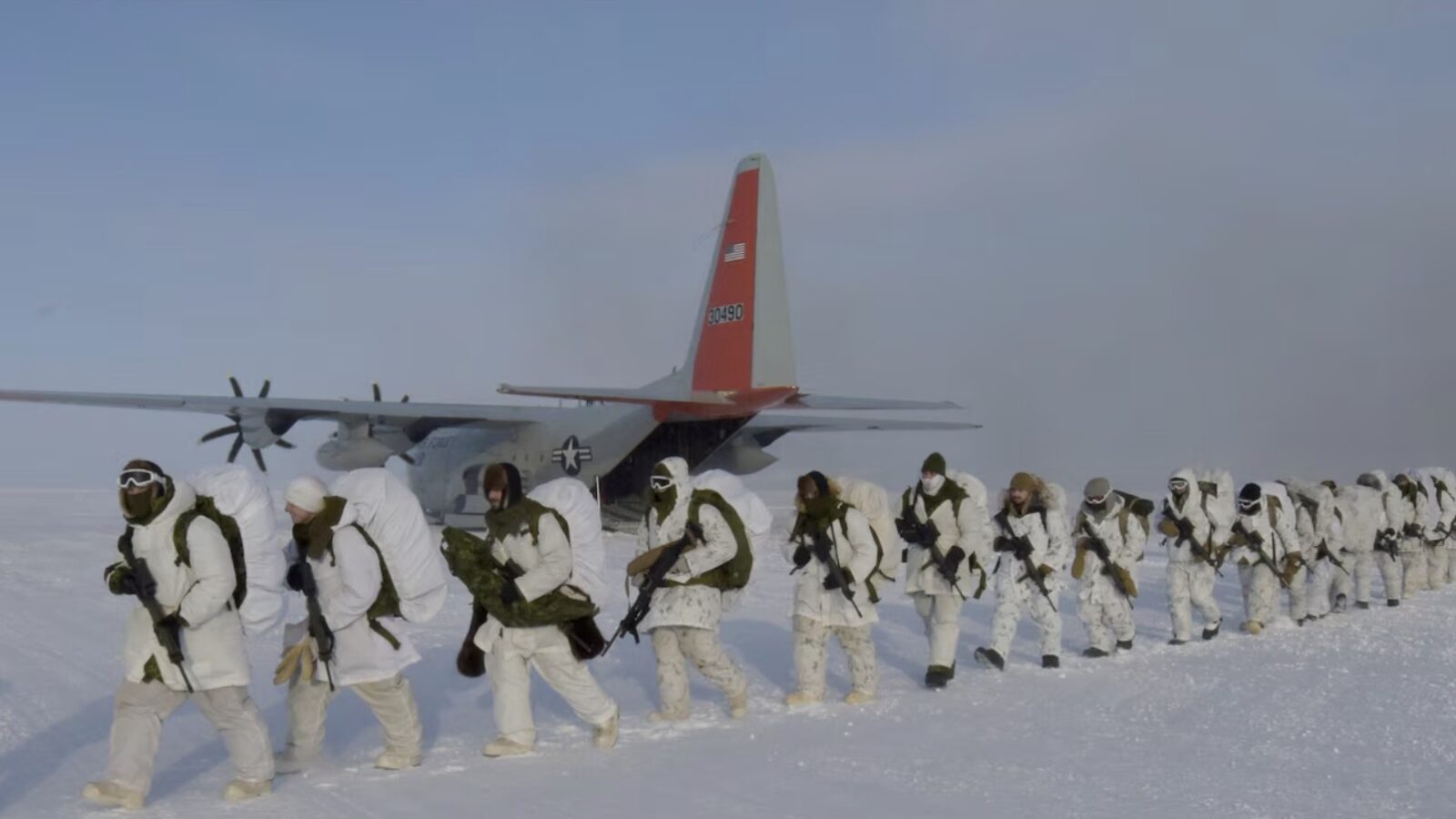 Soldiers in winter gear march across a snowy landscape near a military aircraft, preparing for a cold-weather training exercise.