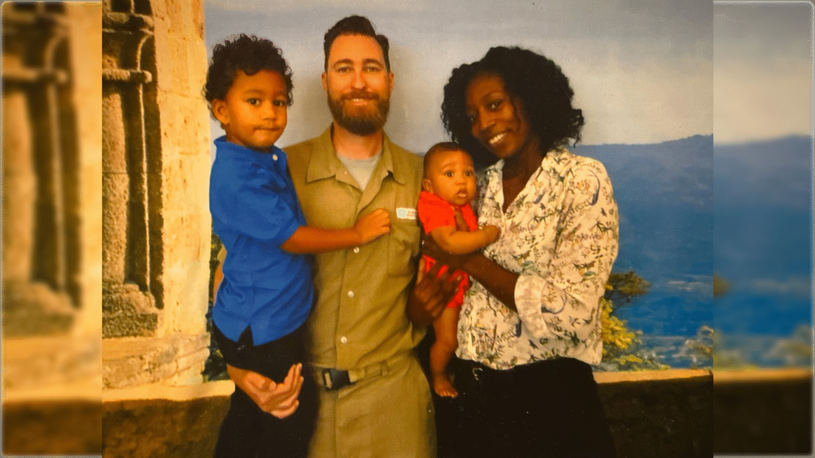 Family portrait featuring a smiling man in a khaki outfit holding a toddler, alongside a woman and an infant, set against a scenic backdrop.