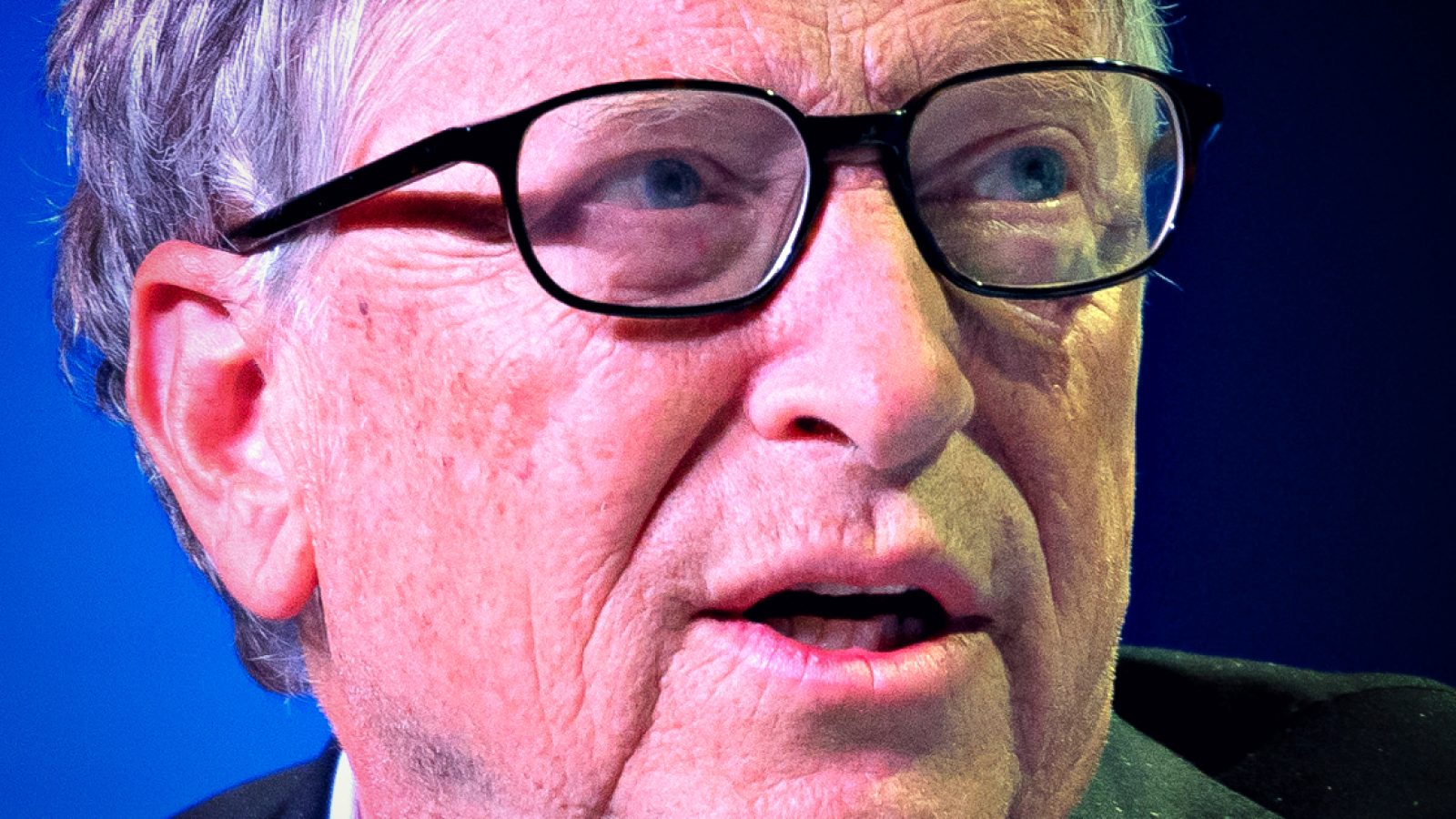 Close-up of a middle-aged man with glasses speaking at an event, showcasing facial expressions and details against a blue background.